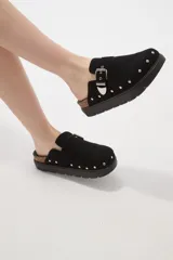 Suecos negros de estilo casual con acabado en eco cuero, detalle de hebilla metálica ajustable en el empeine y tachas decorativas en el contorno de la suela. Presentan una base de corcho y suela de goma dentada.