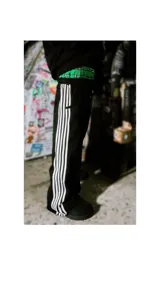 Pantalón deportivo negro de corte ancho, con tres franjas blancas verticales a lo largo de las piernas. La cintura es elástica y presenta un forro interior con estampado a cuadros en tonos verde y negro.