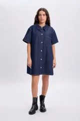 Vestido corto de jean azul oscuro, de manga corta, con cuello camisero, cierre frontal de botones metálicos y un bolsillo aplicado en el pecho.