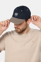 Gorro con visera de lona azul marino y visera en tono tostado. Cuenta con un parche tejido en el frente y ojalillos bordados contrastantes.