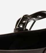 Zapatilla tipo bailarina color marrón oscuro, confeccionada en poliuretano con acabado mate. Presenta punta redondeada, suela plana y dos tiras ajustables con hebillas metálicas plateadas y ojales decorativos sobre el empeine.