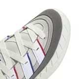 Zapatilla Adidas Adimatic de estilo skate, color gris claro con detalles en gris oscuro. Presenta tres franjas laterales en blanco, con ribetes finos en rojo y azul. La suela es gruesa y texturizada en la puntera.