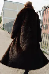 Abrigo largo de piel sintética en color marrón, con cuello de solapa, manga larga y cierre frontal con ganchos metálicos ocultos.