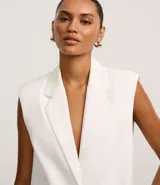 Chaleco largo de sastrería color blanco, con solapas tipo peaked, cierre frontal oculto y ajuste mediante lazo con hebilla en la espalda.