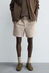 Bermuda de hombre, corte relajado, color beige con estampado de cuadros en tonos marrones y rosados. Presenta cintura elástica, bolsillos frontales y un bolsillo trasero de plastrón con cierre de botón.