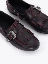 Zapato tipo mocasín de cuero vacuno con efecto gastado y detalles en color rojizo. Presenta flecos en la capellada, una hebilla metálica decorativa y herrajes plateados a lo largo del lateral.