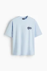 Remera de algodón color celeste con cuello redondo acanalado y hombros caídos. Presenta un estampado gráfico pequeño en el pecho con tipografía estilo graffiti.