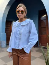 Camisa blanca de corte holgado con cuello bebé con volados, cierre frontal de botones y mangas largas con puños terminados en volados.