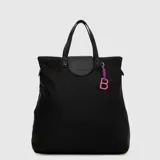 Mochila convertible a bolso tipo tote, color negro, con acabado impermeable. Presenta correas de mano, solapa superior con logo grabado y un llavero decorativo con la letra B en color rosa y violeta.