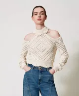 Sweater color crema de punto calado con motivo de rombos, cuello alto con volados y hombros descubiertos. Tiene mangas largas holgadas, ribetes acanalados, bajos festoneados y corte holgado.