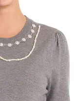 Buzo de tejido fino color blanco, con escote redondo decorado con un collar fijo de perlas y strass. Presenta mangas largas con un leve frunce en los hombros y puños acanalados.