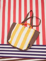 Bolso tipo tote con diseño de rayas verticales en amarillo y blanco, con laterales, base y correas en color marrón. Incluye asas cortas en color naranja y cierre con botón a presión.