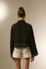 Campera cropped de jean color negro, con cuello camisero, cierre frontal y detalles de tachas plateadas en el cuello, hombros, mangas y ruedo. Presenta un diseño de tachas en forma de corazón en el pecho.