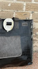 Tote bag de ecocuero negro con textura croco, cuenta con un bolsillo frontal de gamuza marrón y un detalle decorativo circular de guampa en el cierre superior. Posee doble asa de hombro y costuras visibles en los bordes.