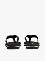 Ojotas negras con tiras de textil trenzado que presentan el logo de Tommy Hilfiger en jacquard con detalles en blanco y dorado. La suela es de EVA dentada con una entresuela a contraste blanca.