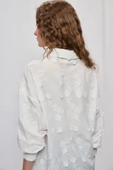 Vestido camisero corto y de silueta amplia en color blanco, con cuello clásico, cierre frontal de botones y mangas largas. Cuenta con diseño bordado floral en toda la prenda y dobladillo asimétrico, más largo en la parte trasera.