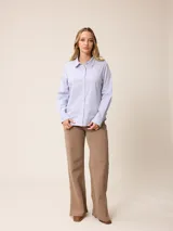 Camisa de corte clásico y calce entallado, color blanco, con cuello camisero, cierre frontal de botones y mangas largas con puños abotonados. Presenta un pequeño bordado distintivo en el pecho.