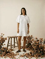 Vestido corto blanco de lino, con cuello redondo con detalle de fruncido y mangas cortas abullonadas. El calce es holgado tipo evasé con un pequeño volante en el ruedo.