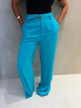 Pantalón de corte amplio color turquesa, de tiro alto con cintura definida y trabillas, acompañado por un cinturón fino a juego con hebilla metálica dorada grabada. Posee bolsillos laterales y pinzas frontales.