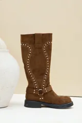Bota alta de gamuza color marrón, con suela plana y gruesa, puntera redondeada y caña alta decorada con costuras en relieve y tachas plateadas. Cuenta con una tira con hebilla y argolla metálica en la base del empeine.