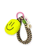 Llavero con aro rosado, dije de peluche amarillo con cara sonriente, cordón trenzado con estampado de pata de gallo, cordón trenzado verde y dije con forma de etiqueta con la marca She Mona y una perla sintética.