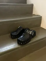 Zuecos negros de cuero encerado con detalle de tachas plateadas en la base y hebilla metálica en el empeine.