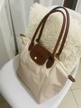 Bolso tipo shopper de cuerpo plegable en color crema o blanco roto, con asas y solapa de cierre en cuero marrón texturizado. Presenta un bolsillo exterior frontal y cierre principal con broche metálico.