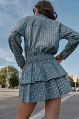 Vestido corto de manga larga con cuello solapa y cierre frontal de botones a presión. Presenta un diseño a cuadros en tonos azul y marrón, cintura ajustable con cinturón de hebilla y falda con volantes en el bajo.