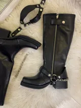 Bota alta tipo biker de cuero negro con cierre metálico lateral que recorre toda la caña. Presenta un diseño versátil con estribo y cadena metálica desmontables en la zona del tobillo, taco cuadrado y plataforma delantera.