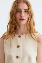 Chaleco color beige con cuello redondo y cierre frontal con botones carey. Presenta diseño forrado y abertura en el dobladillo inferior.