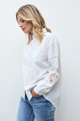 Camisa blanca de corte recto con cuello clásico y cierre frontal de botones. Presenta mangas largas con detalles de encaje calado en los antebrazos y puños abotonados.