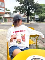 Buzo de manga larga color blanco, corte oversize con hombros caídos. Estampado frontal con la palabra "appetizer" en rojo y una ilustración de un cóctel con aceitunas y rodaja de naranja, acompañado de pequeños cuencos con snacks.