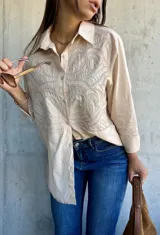 Camisa beige de corte oversize, con cuello clásico y cierre frontal de botones. Presenta un diseño bordado detallado en la parte superior delantera, con mangas largas y un estilo versátil.