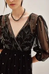 Vestido midi de tul negro con bordados brillantes y pedrería en el escote. Presenta mangas abullonadas, corte imperio y falda con volado en el ruedo. Incluye viso interior.