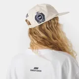 Gorra color beige con visera plana y parche bordado en el frente con escudo y la inscripción 'GOLF'.