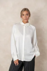 Blusa de satén color marfil con brillo sutil, cuello clásico, cierre frontal con botones y mangas largas con puños abotonados. Presenta un corte fluido y elegante.