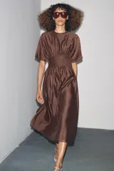 Vestido midi color marrón con textura brillante y arrugada, cuello redondo y mangas cortas tipo murciélago. Presenta un canesú fruncido con pliegues verticales en la cintura que da paso a una falda de corte A.