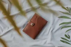 Sobre de cuero color marrón, con cierre de botón a presión en solapa. Presenta costuras visibles en los bordes y el logo grabado en la solapa.