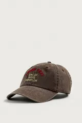 Gorra de seis paneles marrón con efecto lavado, visera curva y texto bordado en relieve en la parte frontal en tonos rojo y amarillo.