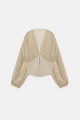 Top beige de manga larga con escote en pico, mangas abullonadas y puños elásticos. Presenta un diseño combinado con tejido fluido en la parte superior y tejido estructurado tipo corsé en la cintura, con cierre frontal de ganchos metálicos.