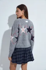 Cardigan corto tejido en color gris, con escote en V y cierre frontal de botones. Presenta un diseño estampado con estrellas y líneas geométricas en tonos oscuros y rojizos, con mangas largas y puños acanalados.