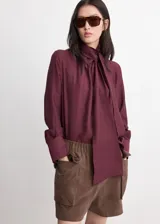 Blusa de manga larga bordó con cuello alto y detalle de fular integrado para anudar al cuello, corte recto y puños abotonados.