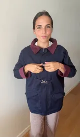 Campera azul marino con cuello y puños en color bordó, diseño con cierre frontal y broches metálicos tipo gancho, y bolsillos delanteros con solapa.