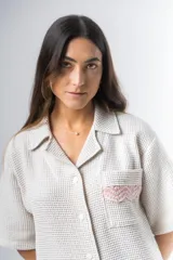 Conjunto de pijama de tela waffle color crema, compuesto por una camisa de manga corta con botones, bolsillo y detalle de broderie, y un short con elástico y cordón en la cintura con terminación de broderie.