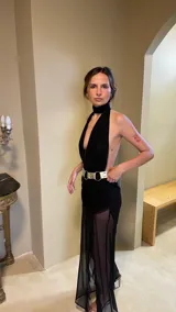 Vestido largo de color negro confeccionado en microtul. Presenta un escote profundo tipo halter con cuello alto, falda con frunces en la zona de la cadera y transparencias en la parte inferior.