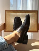 Mocasín de cuero color chocolate con acabado texturizado, diseño de punta cuadrada y suela plana con pequeño tacón.
