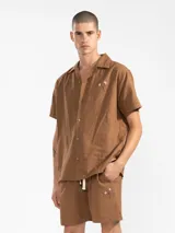 Bermuda de hombre color azul petróleo, confeccionada en mezcla de lino y algodón. Presenta cintura elástica con cordón, bolsillos laterales y trasero tipo parche. Incluye bordado floral en la pierna izquierda y etiqueta con logo en el bolsillo trasero.