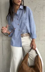 Camisa clásica de manga larga, a rayas verticales azules y blancas, con cuello camisero y botones frontales. Presenta un bordado lineal de un rostro en el frente.