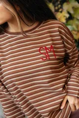Remerón de manga larga con diseño de rayas horizontales finas en tonos tierra. Presenta un corte holgado y cuenta con un bordado artesanal en color rojo en el pecho.
