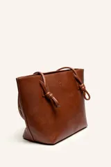 Cartera tote de cuero suave color marrón, con diseño amplio y líneas simples, con tiras de hombro decoradas con nudos artesanales y pequeño logo grabado en el centro.